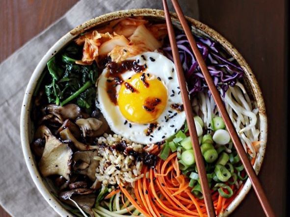 Un colorido tazón de bibimbap con un huevo frito, verduras vibrantes, champiñones y arroz se presenta artísticamente junto con palillos, que recuerdan al Taller de cocina coreana en Sevilla. - ForTwo - Planes para parejas