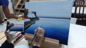 En un estudio sereno, un artista se inspira en Bibiana Suárez durante un taller creativo, utilizando técnicas de "Talleres Creativos con Bibiana Suárez: Aprende Técnicas Multidisciplinarias" para transformar una fotografía en un vibrante paisaje costero con edificios bajo un cielo tranquilo. - ForTwo - Planes para parejas