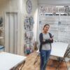 Una persona sonriendo en una sala de manualidades con suministros organizados, mesas y un gran reloj de pared, que recuerda a "Talleres Creativos con Bibiana Suárez: Aprende Técnicas Multidisciplinarias". - ForTwo - Planes para parejas