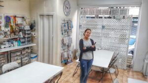 Una persona sonriendo en una sala de manualidades con suministros organizados, mesas y un gran reloj de pared, que recuerda a "Talleres Creativos con Bibiana Suárez: Aprende Técnicas Multidisciplinarias". - ForTwo - Planes para parejas