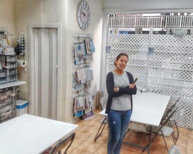 Una persona sonriendo en una sala de manualidades con suministros organizados, mesas y un gran reloj de pared, que recuerda a "Talleres Creativos con Bibiana Suárez: Aprende Técnicas Multidisciplinarias". - ForTwo - Planes para parejas