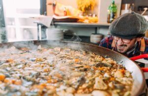 En un acogedor rincón del Espai Granada, una persona con gafas y gorra admira una gran cacerola humeante de guiso de verduras mixtas, parecido a los deliciosos 'Talleres de cocina con arroces en Espai Granada'. - ForTwo - Planes para parejas