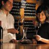 Dos personas disfrutan de un vino tinto en una barra de madera en el ambiente acogedor de "Visita a una Bodega Tradicional en Granada: Un paseo por los secretos del vino". Con decantador, copas y estanterías repletas de vinos locales, cada sorbo cuenta una historia de tradición y sabor. - ForTwo - Planes para parejas