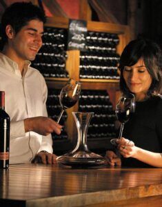 Dos personas disfrutan de un vino tinto en una barra de madera en el ambiente acogedor de "Visita a una Bodega Tradicional en Granada: Un paseo por los secretos del vino". Con decantador, copas y estanterías repletas de vinos locales, cada sorbo cuenta una historia de tradición y sabor. - ForTwo - Planes para parejas