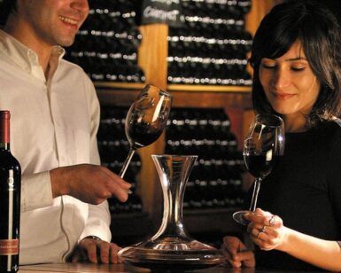 Dos personas disfrutan de un vino tinto en una barra de madera en el ambiente acogedor de "Visita a una Bodega Tradicional en Granada: Un paseo por los secretos del vino". Con decantador, copas y estanterías repletas de vinos locales, cada sorbo cuenta una historia de tradición y sabor. - ForTwo - Planes para parejas