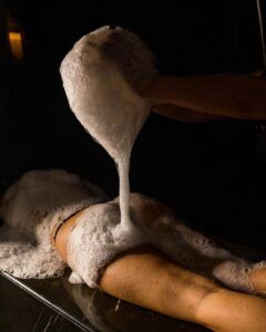 En una sala poco iluminada, una persona se relaja en una mesa, rodeada de relajantes burbujas durante un placentero masaje con espuma, que recuerda la experiencia de lujo que ofrece el Baño Turco con Café y Chocolate en Madrid. - ForTwo - Planes para parejas