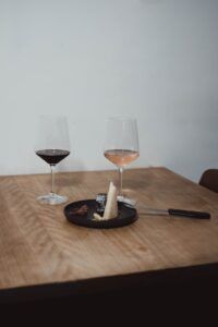 En una mesa de madera en Madrid, dos copas de vino tinto y rosado se combinan con cuñas de queso y un cuchillo en un plato negro: una cata de quesos con maridaje perfecta. - ForTwo - Planes para parejas