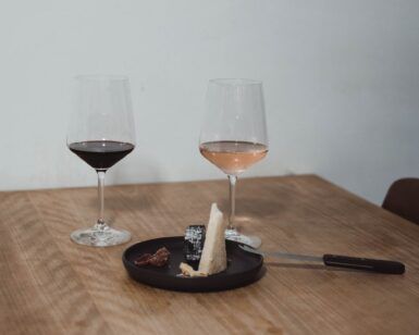 En una mesa de madera en Madrid, dos copas de vino tinto y rosado se combinan con cuñas de queso y un cuchillo en un plato negro: una cata de quesos con maridaje perfecta. - ForTwo - Planes para parejas