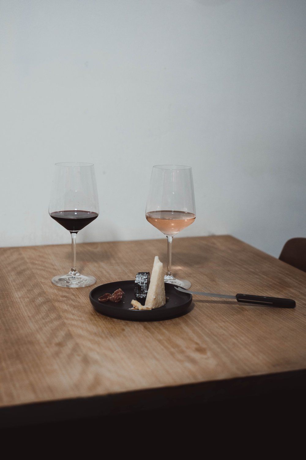 En una mesa de madera en Madrid, dos copas de vino tinto y rosado se combinan con cuñas de queso y un cuchillo en un plato negro: una cata de quesos con maridaje perfecta. - ForTwo - Planes para parejas