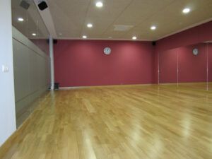 El estudio de baile vacío, con suelo de madera y paredes rojas, es ideal para clases de baile de salón en Madrid: ritmos latinos, swing y tango. Grandes espejos y luces de techo crean un ambiente vibrante, con un reloj de pared que garantiza movimientos apasionados y puntuales. - ForTwo - Planes para parejas