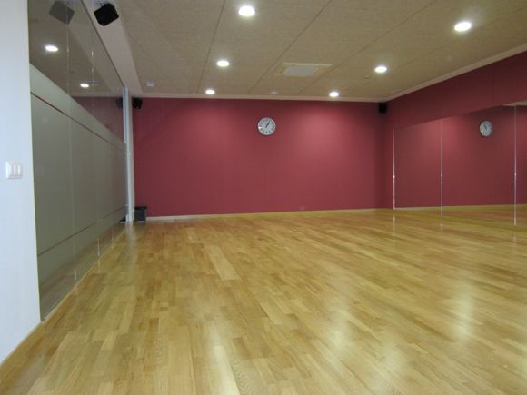 El estudio de baile vacío, con suelo de madera y paredes rojas, es ideal para clases de baile de salón en Madrid: ritmos latinos, swing y tango. Grandes espejos y luces de techo crean un ambiente vibrante, con un reloj de pared que garantiza movimientos apasionados y puntuales. - ForTwo - Planes para parejas