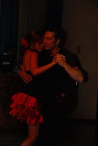 Bajo la íntima luz tenue, una pareja baila muy cerca en Clases de Bailes de Salón en Madrid, con la mujer vestida de rojo y el hombre de negro. Se mueven con gracia, sus pasos susurran historias de veladas apasionadas. - ForTwo - Planes para parejas