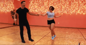 Un hombre y una mujer bailan con gracia en una habitación con paredes naranjas y luces de colores, evocando el encanto de las clases de baile de salón en Madrid. - ForTwo - Planes para parejas