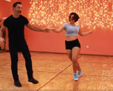 Un hombre y una mujer bailan con gracia en una habitación con paredes naranjas y luces de colores, evocando el encanto de las clases de baile de salón en Madrid. - ForTwo - Planes para parejas