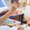 Primer plano de alguien que asiste a Clases de Dibujo y Pintura en Zaragoza, pintando sobre un lienzo con un pincel y sosteniendo una paleta llena de colores vibrantes. - ForTwo - Planes para parejas