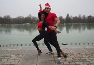 Dos personas con camisas rojas y sombreros festivos bailan alegremente salsa cubana junto a un lago en un día nublado, exudando una energía vibrante que recuerda a las clases de salsa cubana en Madrid. - ForTwo - Planes para parejas