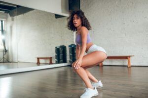 En un estudio de Madrid, una persona posa con confianza con ropa deportiva frente a un gran espejo y aparatos de ejercicio, lista para su sesión de Clases de Twerk en Madrid. - ForTwo - Planes para parejas