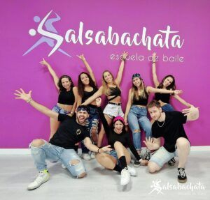 Un grupo de personas sonrientes posan enérgicamente frente a una pared violeta vibrante que dice Clases de Twerk en Madrid, emocionadas después de su animada sesión de baile. - ForTwo - Planes para parejas