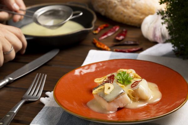 Un plato de pescado en un plato naranja, cubierto con salsa y láminas de ajo, invita a disfrutar de la diversión culinaria. Rodeado de cubiertos y especias sobre una mesa de madera, captura la magia de la cocina, como en "Divídete Cocinando en Madrid A Punto". - ForTwo - Planes para parejas