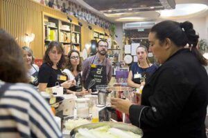 En una cocina madrileña bien iluminada, un chef vestido de negro dirige una atenta clase de cocina, capturando a la perfección la esencia de "Diviértete Cocinando en Madrid A Punto". - ForTwo - Planes para parejas