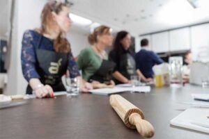Un rodillo reposa sobre la encimera de la cocina mientras en el fondo se ven personas con delantales, posiblemente de la aclamada película "Diviértete cocinando en Madrid a punto". - ForTwo - Planes para parejas