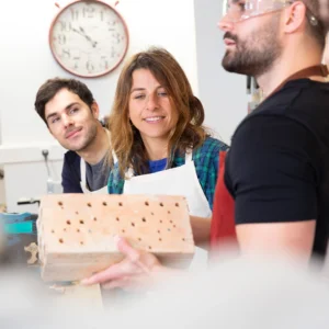 Tres personas se encuentran en el Taller de Bricolaje de Supervivencia de Barcelona; dos observan mientras una sostiene un bloque perforado. Un reloj de fondo les recuerda sutilmente la hora de Barcelona. - ForTwo - Planes para parejas