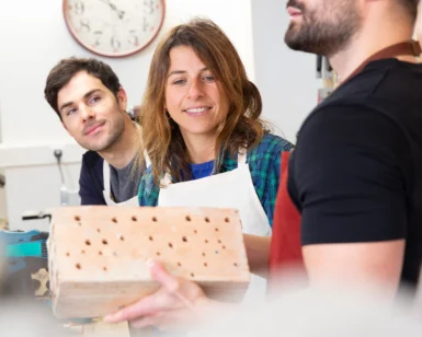 Tres personas se encuentran en el Taller de Bricolaje de Supervivencia de Barcelona; dos observan mientras una sostiene un bloque perforado. Un reloj de fondo les recuerda sutilmente la hora de Barcelona. - ForTwo - Planes para parejas