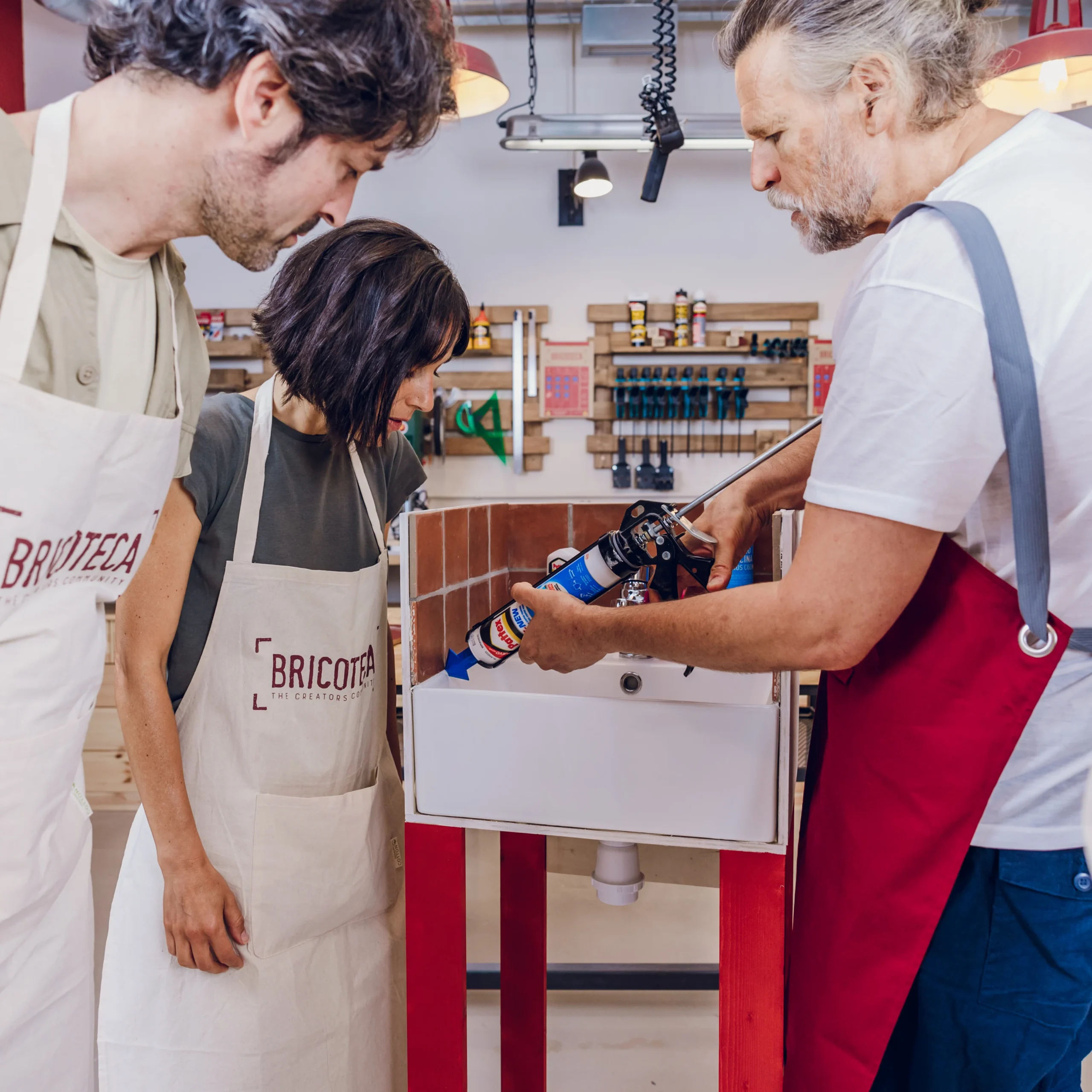 En un taller, tres personas con delantales colaboran en un proyecto de fregadero, convirtiendo sus esfuerzos en un atractivo Taller de Bricolaje de Supervivencia en Barcelona, donde las habilidades y la creatividad florecen entre las herramientas como en un rincón artesano. - ForTwo - Planes para parejas