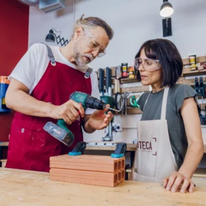 En un concurrido Taller de Bricolaje de Supervivencia en Barcelona, dos personas trabajaban diligentemente en su proyecto, usando un taladro sobre ladrillos. Con gafas de seguridad y delantales, encarnaban el espíritu de supervivencia mediante el desarrollo práctico de habilidades. - ForTwo - Planes para parejas