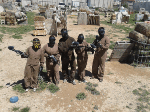Cinco personas con monos marrones y máscaras viven una emocionante aventura de paintball en un campo al aire libre con ¡Acción y Diversión en Paintballmania Zaragoza!, lanzándose entre fardos de heno y desatando vibrantes explosiones de color para una experiencia llena de emoción. - ForTwo - Planes para parejas