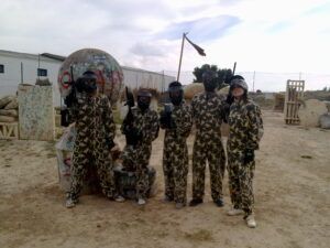 Cinco personas con equipo de camuflaje y máscaras sostienen pistolas de paintball en un campo al aire libre, listas para la acción en ¡Acción y Diversión en Paintballmania Zaragoza!, donde la diversión se encuentra con la estrategia. - ForTwo - Planes para parejas