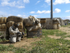 Agachado detrás de fardos de heno en un campo de hierba bajo un cielo azul, alguien vestido para la acción se prepara para disfrutar de ¡Acción y Diversión en Paintballmania Zaragoza!. - ForTwo - Planes para parejas