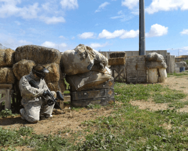 Agachado detrás de fardos de heno en un campo de hierba bajo un cielo azul, alguien vestido para la acción se prepara para disfrutar de ¡Acción y Diversión en Paintballmania Zaragoza!. - ForTwo - Planes para parejas