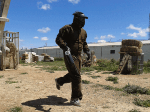 Con equipo de protección oscuro y máscara, una persona camina por un campo de tierra entre fardos de heno cerca de un almacén, encarnando la emoción de ¡Acción y Diversión en Paintballmania Zaragoza!. - ForTwo - Planes para parejas