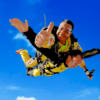 Dos personas disfrutan de un emocionante Salto Tándem en Paracaídas, saltando contra un cielo azul vibrante, mientras una de ellas levanta el pulgar. - ForTwo - Planes para parejas