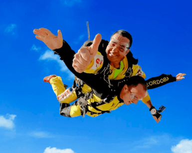 Dos personas disfrutan de un emocionante Salto Tándem en Paracaídas, saltando contra un cielo azul vibrante, mientras una de ellas levanta el pulgar. - ForTwo - Planes para parejas
