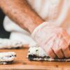 Una persona con guantes enrolla sushi con destreza sobre una tabla de madera, perfeccionando su oficio con técnicas del "Taller de Cocina Japonesa: Aprende a Preparar Sushi, Gyozas y Más". - ForTwo - Planes para parejas