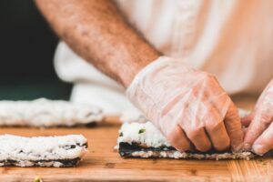 Una persona con guantes enrolla sushi con destreza sobre una tabla de madera, perfeccionando su oficio con técnicas del "Taller de Cocina Japonesa: Aprende a Preparar Sushi, Gyozas y Más". - ForTwo - Planes para parejas