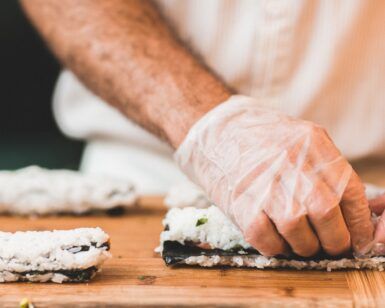 Una persona con guantes enrolla sushi con destreza sobre una tabla de madera, perfeccionando su oficio con técnicas del "Taller de Cocina Japonesa: Aprende a Preparar Sushi, Gyozas y Más". - ForTwo - Planes para parejas