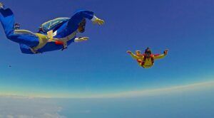Contra un cielo azul claro, dos paracaidistas vestidos con trajes azules y amarillos disfrutan de la emoción de un Salto Tándem en Paracaídas con sus paracaídas abiertos. - ForTwo - Planes para parejas