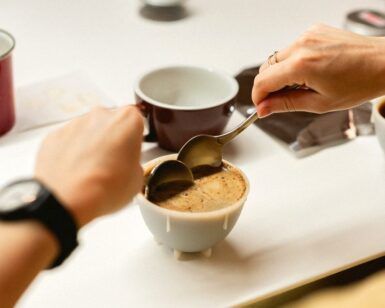 Las manos sostienen cucharas sobre el café, saboreando el momento, mientras las tazas cercanas insinúan una deliciosa experiencia de "Cata de Café en Cafés el Magnífico: Aprende a Valorar los Matices del Café. - ForTwo - Planes para parejas
