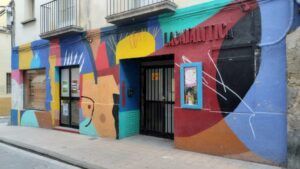 Un vibrante mural adorna la fachada de un edificio en Huesca con formas geométricas abstractas, con el texto "La Carátula" sobre la puerta. Esto invita a los transeúntes a sumergirse en su encanto, quizás inspirándolos a tomar "Aprende a Bailar Swing en Huesca: Clases de Lindy Hop para Todos los Niveles" en las inmediaciones. - ForTwo - Planes para parejas