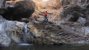 Dos aventureros con trajes de neopreno y cascos saltan a una poza rocosa debajo de una pequeña cascada, mostrando la emoción del "Barranquismo en Los Carrizales, Tenerife: Aventura Extrema. - ForTwo - Planes para parejas