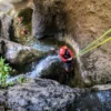 En las gargantas rocosas de Tenerife, una persona con casco y traje de neopreno desciende por una cascada durante la emocionante aventura "Barranquismo en Los Carrizales, Tenerife: Aventura Extrema", mientras otro escalador espera abajo. - ForTwo - Planes para parejas