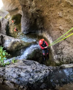 En las gargantas rocosas de Tenerife, una persona con casco y traje de neopreno desciende por una cascada durante la emocionante aventura "Barranquismo en Los Carrizales, Tenerife: Aventura Extrema", mientras otro escalador espera abajo. - ForTwo - Planes para parejas