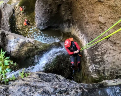 En las gargantas rocosas de Tenerife, una persona con casco y traje de neopreno desciende por una cascada durante la emocionante aventura "Barranquismo en Los Carrizales, Tenerife: Aventura Extrema", mientras otro escalador espera abajo. - ForTwo - Planes para parejas