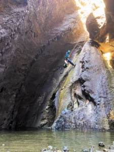 Una persona desciende en rápel por un acantilado rocoso dentro de una enorme cueva en Tenerife, con el agua brillando debajo y la luz del sol entrando desde arriba, capturando la esencia del Barranquismo en Los Carrizales, Tenerife: Aventura Extrema. - ForTwo - Planes para parejas