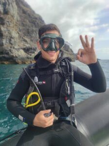 Un buzo con traje de neopreno y máscara da la señal de OK en un barco con los acantilados rocosos de El Hierro detrás, listo para su Bautismo de Buceo en El Hierro: Vive tu Primera Inmersión. - ForTwo - Planes para parejas