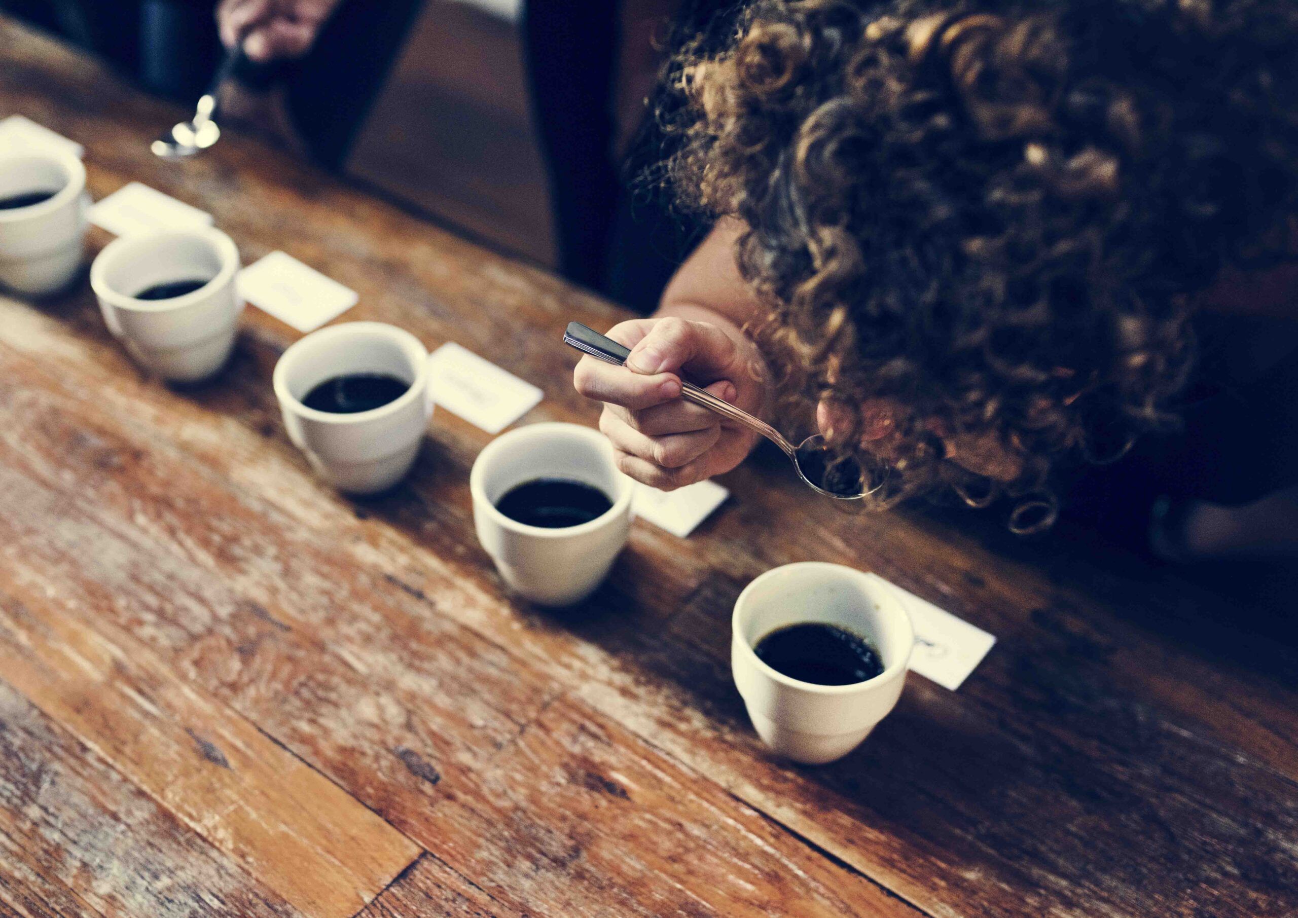 Una persona participa en la Cata Sensorial de Café, probando ricos sabores y fragancias de múltiples tazas en una mesa de madera, usando una cuchara para saborear cada nota. - ForTwo - Planes para parejas