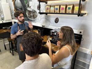 En la acogedora cafetería, el barista describió con pasión "Cata Sensorial de Café: Descubre Fragancias, Sabores y Cata Ciega" a dos clientes, enriqueciendo su apreciación con palabras sobre experiencias sensoriales en medio de ricos aromas. Cerca de allí, las bolsas de café en un estante prometían una infinidad de sabores para explorar. - ForTwo - Planes para parejas